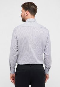 Witte herenoverhemd met blauw geruit patroon. Lange mouwen met manchetknopen, klassieke kraag en een getailleerde pasvorm. Zwarte broek zichtbaar.