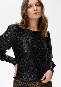 Vrouw in een zwart glinsterend top met lange mouwen en broek met luipaardprint, poserend met één hand op haar heup en de andere bij haar taille.