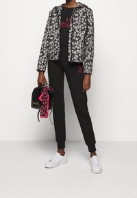 Blommönstrad svart jacka, svart topp med röd text, matchande joggers, vita sneakers och en kviltad svart väska med rosa scarf-accent.