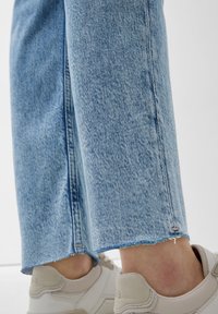 Mezclilla azul claro con dobladillos sin rematar, combinada con zapatillas de deporte blanco roto, sobre un fondo blanco.