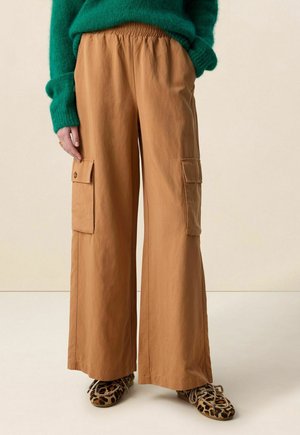 Personne portant un pantalon cargo large couleur sable avec des poches latérales à boutons, un pull vert en matière pelucheuse et des baskets à imprimé léopard.
