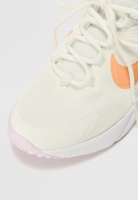 Zapatilla deportiva de color claro con un upper texturizado, que presenta un logo de swoosh naranja, punta redondeada y una suela con tintes rosas.