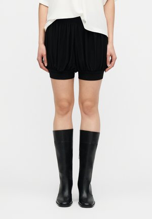 Nő fekete, ráncolt sortot, fehér felsőt és térdig érő fekete bőr csizmát visel, karjai laza tartásban oldalán áll.