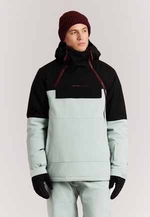 Man draagt een zwarte en lichtblauwe winterjas met capuchon, zwarte handschoenen, lichtblauwe broek en een donkerrode gebreide muts, staand tegen een effen achtergrond.