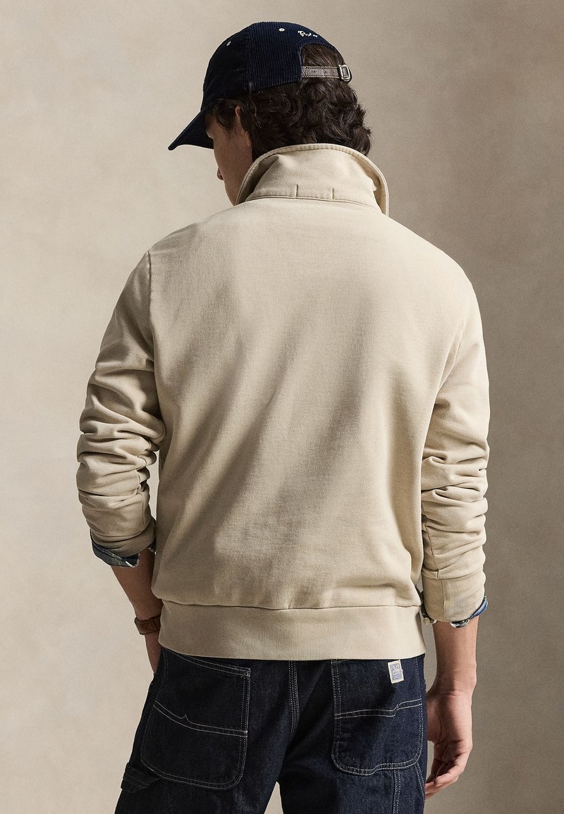 Man wearing beige pullover and navy cap, hands in pockets, viewed from behind against a neutral background.