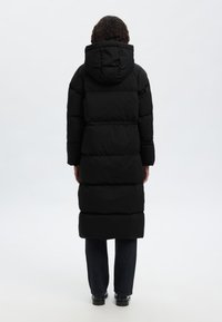 Zwarte gewatteerde lange jas met capuchon, met horizontale stiksels, een getailleerde taille en zijzakken. Draagt donkere jeans en zwarte schoenen.