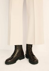 Ciemnobrązowe skórzane buty z wytłoczeniem w kształcie krokodyla, ze złotymi oczkami, czarną gumową podeszwą i sznurowanym designem, w połączeniu z kremowymi spodniami o szerokich nogawkach.