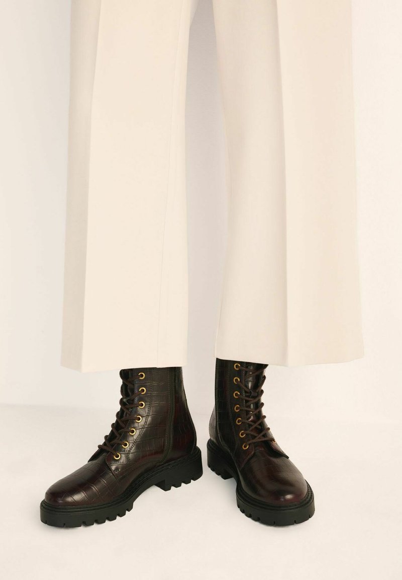 Ciemnobrązowe skórzane buty z wytłoczeniem w kształcie krokodyla, ze złotymi oczkami, czarną gumową podeszwą i sznurowanym designem, w połączeniu z kremowymi spodniami o szerokich nogawkach.