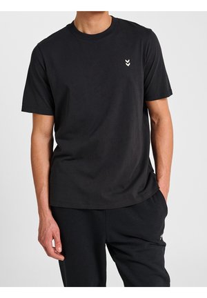 Homme portant un t-shirt noir uni à manches courtes avec un petit logo double chevron blanc en haut à gauche et un pantalon noir.