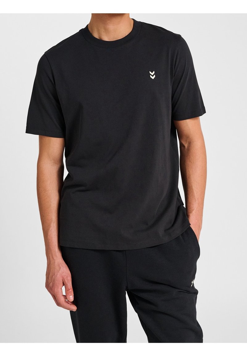 Homme portant un t-shirt noir uni à manches courtes avec un petit logo double chevron blanc en haut à gauche et un pantalon noir.