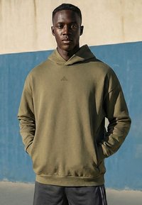 Jeune homme debout en extérieur portant un sweat à capuche vert olive, les mains dans les poches, devant un mur bleu et beige.