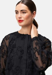Schwarze Bluse mit durchsichtigen langen Ärmeln und strukturiertem floralem Stickereimuster. Rundhalsausschnitt und Knopfverschluss. Glattes Gewebe mit einem subtilen Glanz.