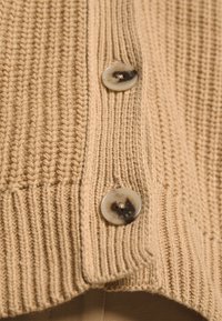 Beige neulejakku, jossa on teksturoitu kuvio, varustettuna kahdella pyöreällä, vaalealla napilla, joissa on logoyksityiskohta etuhalkiossa.