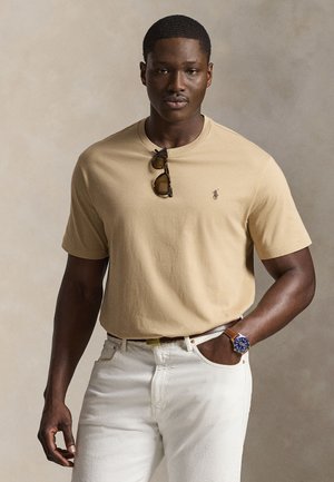 Izmos férfi bézs pólóban, napszemüveggel a gallérján, fehér nadrágban és kék karórával, egy egyszínű bézs háttér előtt áll.