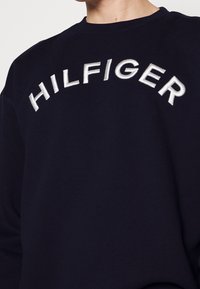 Sudadera azul marino con cuello redondo, que presenta un gran texto bordado en blanco "HILFIGER" en el pecho. Tejido suave, corte casual, con un detalle minimalista.
