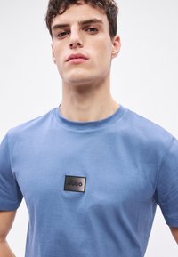 Modré bavlněné tričko s kulatým výstřihem a černou obdélníkovou nášivkou s nápisem „HUGO“ na přední straně. Hladká textura.
