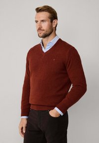 V-Ausschnitt-Pullover in tiefem Rostfarben, aus weichem Strickstoff gefertigt, mit gerippten Bündchen und Saum. Über einem hellblauen Hemd mit Kragen getragen.
