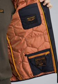 Die Innenausstattung der marineblauen, gesteppte Jacke verfügt über ein Kupferfutter, ein sechseckiges Muster, gelbe Akzente und mehrere Taschen mit Markenetiketten.