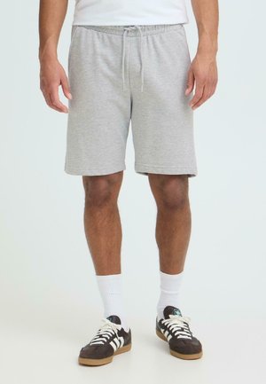 Uomo che indossa pantaloncini grigi chiaro con coulisse, calzini bianchi alla caviglia e scarpe da ginnastica nere con strisce bianche, in piedi davanti a uno sfondo semplice.