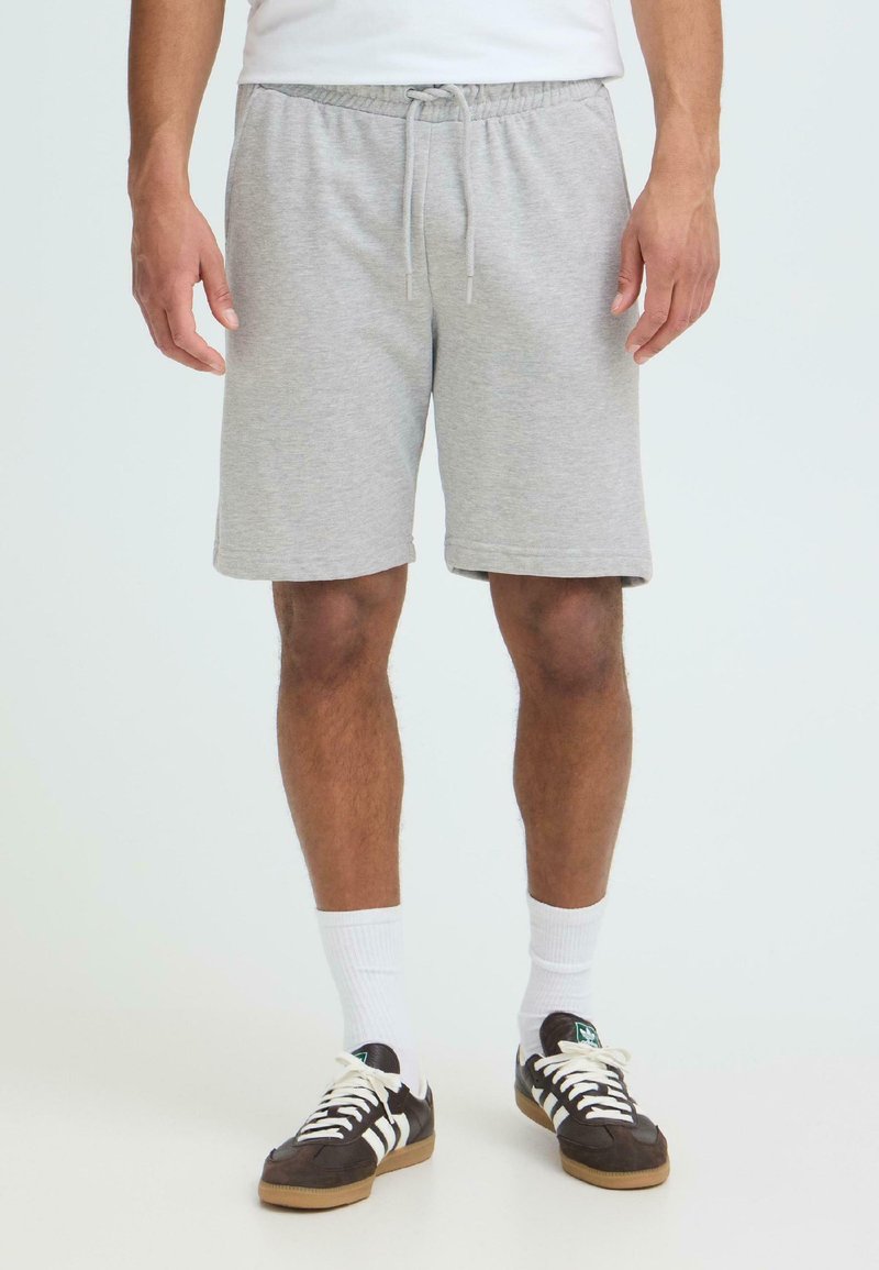 Homme portant un short gris clair à cordon, des chaussettes blanches montantes et des baskets noires à rayures blanches, debout devant un fond uni.