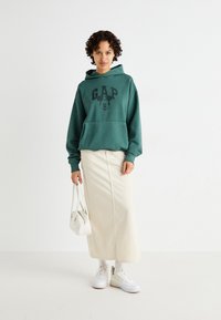 Grønn bomull hettegenser med svart "GAP" logo og Mickey Mouse grafikk, kombinert med en krem midi-skjørt og hvite joggesko, holder en hvit veske.