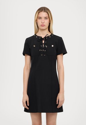 Model in a black dress featuring a lace-up front and two chest pockets, posing against a light background with a neutral expression.