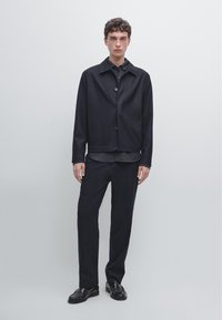 Ciemna kurtka z mieszanki wełny z kołnierzem, zapinana z przodu na guziki, w zestawieniu z ciemną koszulą zapinaną na guziki i dopasowanymi spodniami. Czarne skórzane buty.