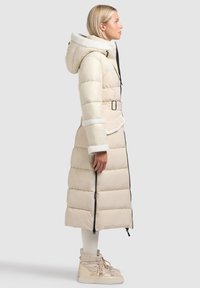 Lange gewatteerde jas in lichtbeige met een met bont gevoerde capuchon, een taille met ceintuur en ritssluitingen aan de zijkanten. Gecombineerd met chunky beige sneakers.