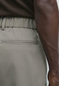 Primo piano di una persona che indossa pantaloni grigi chiari con vita elastica e tasca a filetto sul retro, abbinati a una maglietta marrone scuro.