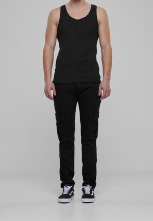 Hombre vistiendo una camiseta sin mangas negra, pantalones cargo negros y zapatillas blancas y negras, de pie sobre un fondo gris.