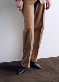 Pantalons taillés couleur beige avec une coupe droite, associés à des talons noirs pointus. Des bagues métalliques ornent les doigts, sur un tapis brun texturé.