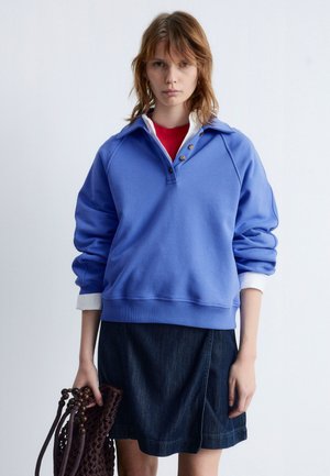 Light blue pullover sweatshirt with a collar, three buttons, and white cuffs, paired with a dark denim skirt. Features a knitted brown bag.