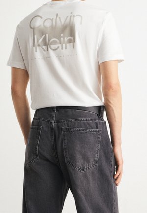 Person wearing a white Calvin Klein T-shirt with logo text on the back and high-waisted black denim jeans, standing facing away.