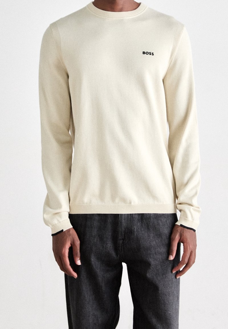 Homme portant un pull à manches longues crème avec le logo « BOSS » sur la poitrine et un pantalon gris foncé sur un fond uni.