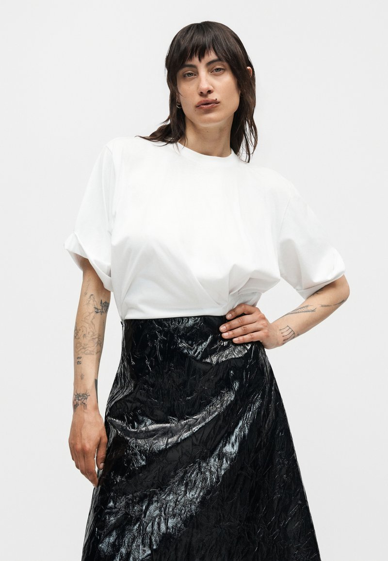 Kvinde med mørkt hår iført en hvid oversized top med korte ærmer og et sort skinnende tekstureret skørt, poserer med den ene hånd på hoften.
