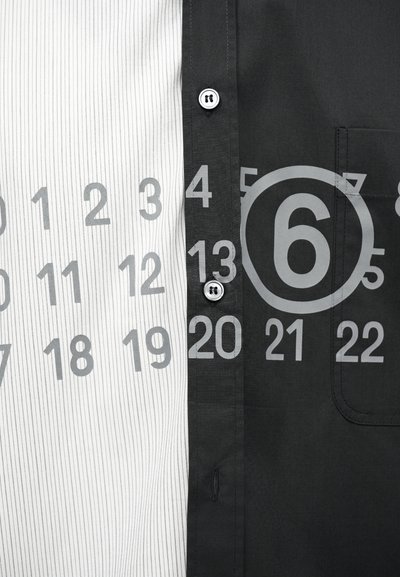 Κοντινή λήψη ενός ασπρόμαυρου πουκαμίσου με γκρι αριθμούς ημερολογίου τυπωμένους σε όλη την επιφάνεια, με έμφαση στον αριθμό 6 που είναι κύκλος στη μαύρη πλευρά.