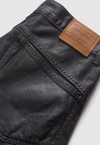 Jeans en denim noir avec un patch en cuir de la marque. Le tissu a un léger éclat, avec des coutures visibles et une texture lisse.