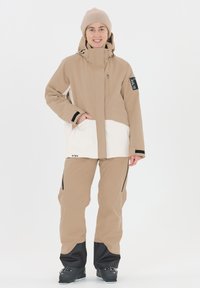 Giacca da sci beige con una parte inferiore bianca e una tasca con zip, abbinata a pantaloni beige con dettagli neri e zip.