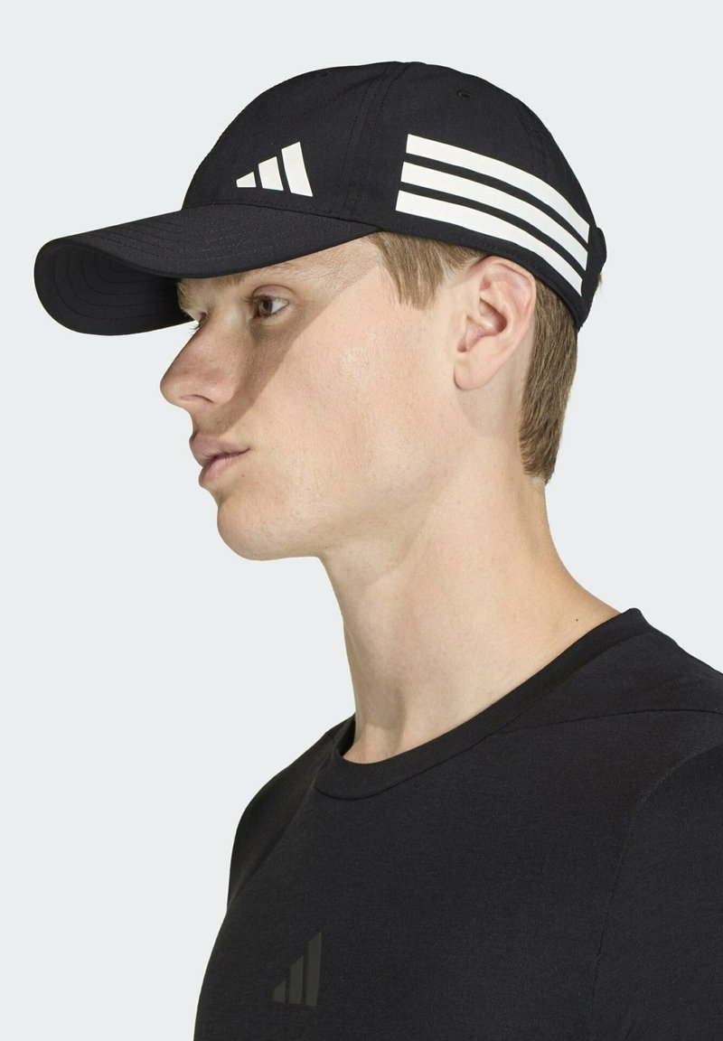 Gorra de béisbol negra con un diseño de tres rayas blancas en el lateral; tiene una visera curvada y una textura suave. Se lleva con una camiseta negra.