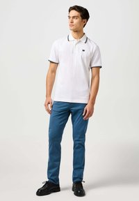 Camisa polo blanca con detalles negros y un pequeño logo, combinada con pantalones azul claro y zapatos negros, con un diseño ajustado.