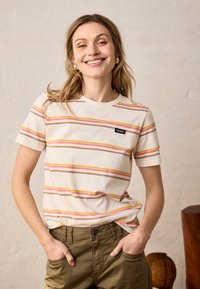 Femme souriante, portant un t-shirt crème à manches courtes avec des rayures roses, jaunes et marron, et un pantalon vert olive les mains dans les poches.