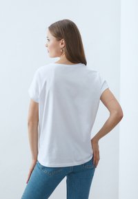 White cotton t-shirt with short sleeves, round neckline, and a relaxed fit, paired with light blue jeans. Back view shows simple design.