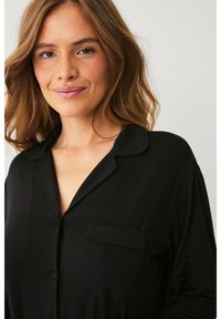 Chemise noire à boutons avec une texture douce, un col et une poche simple sur le côté gauche. Tissu léger qui tombe légèrement sur le corps.