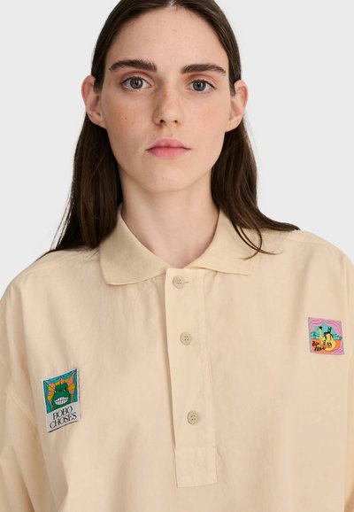 Jeune femme aux longs cheveux bruns porte une chemise beige boutonnée avec des patches colorés sur la poitrine sur fond uni.