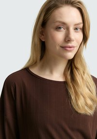Jonge vrouw met lang blond haar, gekleed in een geribbelde bruine top, die iets naar rechts is gekeerd met een vriendelijke glimlach, neutrale lichte achtergrond.