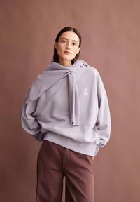 Hoodie oversized lavande avec un logo blanc, doté d'un col rond et de manches longues. Associé à un pantalon marron foncé, sur un fond neutre.
