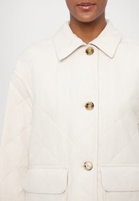 Quilted cream jacket with diamond pattern, large collar, two front pockets, and button closures featuring gold-tone hardware.