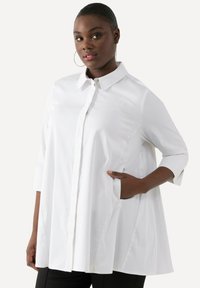 Blusa bianca con colletto a punta, chiusura frontale con bottoni e maniche a tre quarti. Presenta una vestibilità ampia e tasche laterali. Tessuto liscio.