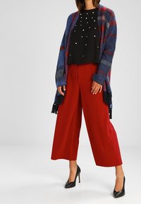 Cardigan di maglia oversize blu con strisce rosse, top nero con dettagli di perle, pantaloni a gamba larga rossi e scarpe con tacco nere.