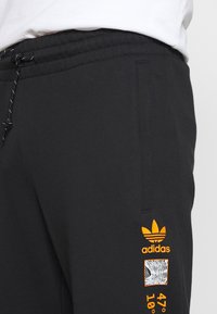 Pantaloni della tuta neri in cotone con vita elastica, decorati con un logo arancione Adidas e grafiche sul lato sinistro.